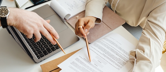 hands holding pencils pointing at paper with a laptop