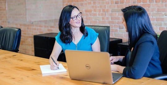 two women sitting down at a table with a laptop and pad of paper, and a brick wall behind them