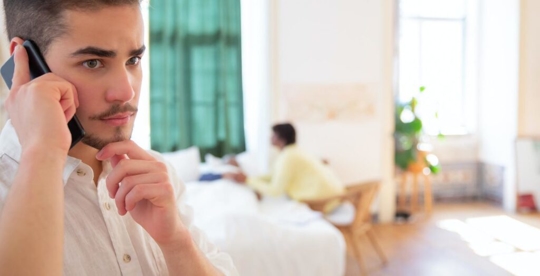 gentleman talking on a cell phone with someone lying in the bed behind him
